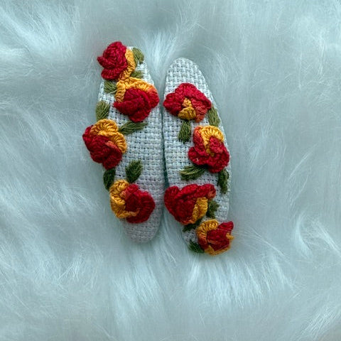 A pair of white knit slip-on slippers decorated with red and yellow crocheted flowers and green leaves, laid flat on a white faux fur surface.