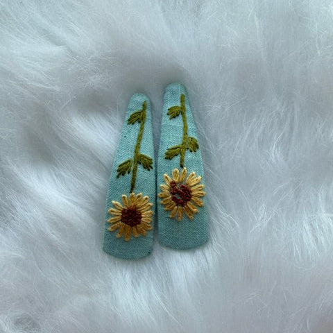 A pair of light blue fabric hair clips decorated with embroidered sunflowers and green stems, displayed on a white faux fur background.