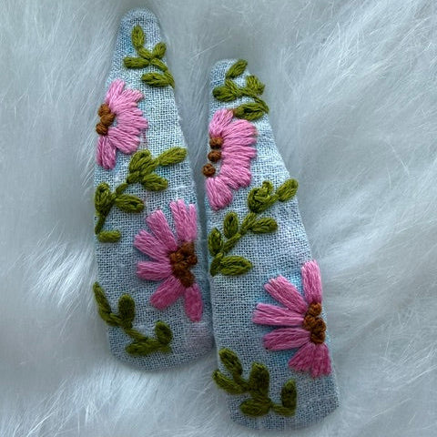 A pair of pointed fabric hair clips decorated with pink embroidered flowers and green leaves on a light gray background, displayed on a white faux fur surface.