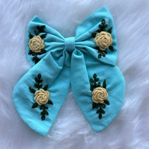 A light blue fabric hair bow decorated with three beige embroidered roses and dark green leaves, displayed on a white faux fur background.