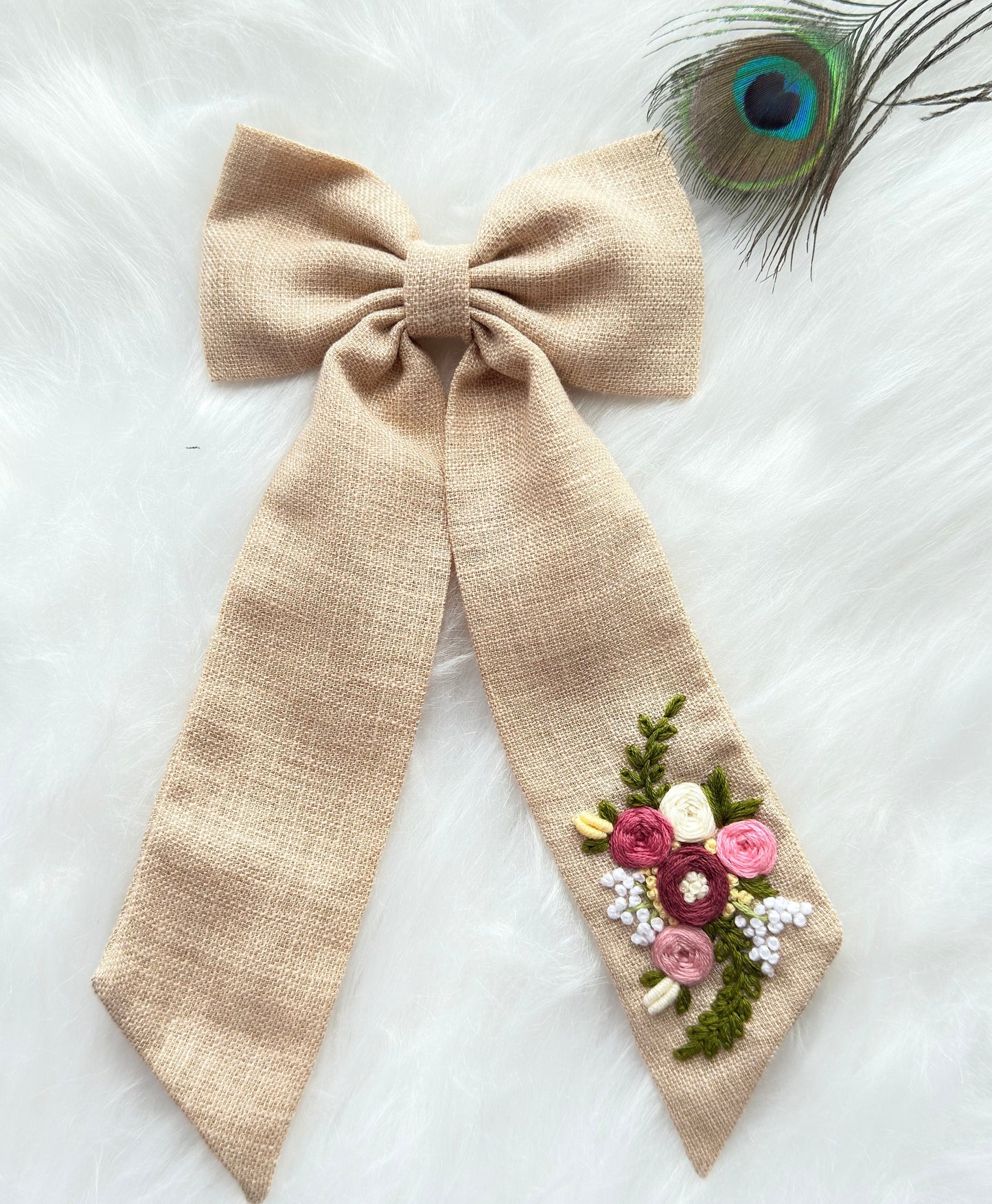 A beige linen ribbon bow with a long tail decorated with a small embroidered floral cluster in pink, burgundy, green, and white, displayed on a white faux fur background with a peacock feather accent.